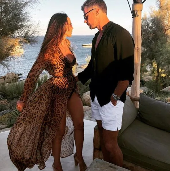 Couple enjoying a vacation by the sea, standing on a patio with scenic ocean and trees in the background.