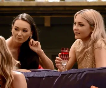 Two women seated on a couch, one holding a red glass, engaged in conversation.