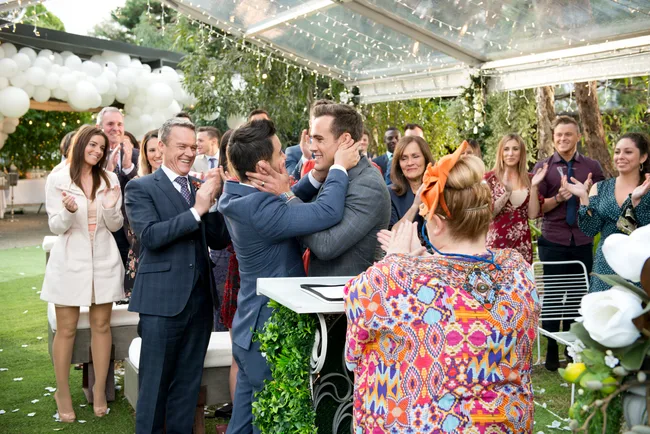A wedding scene with two men embracing and guests applauding under a canopy decorated with balloons and lights.