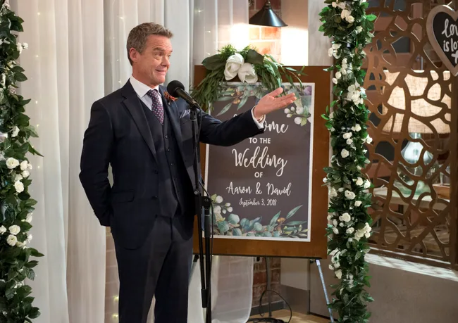 Wedding scene with a man in a suit speaking at a microphone, floral decor, and a "Welcome to the Wedding of Aaron & David" sign.