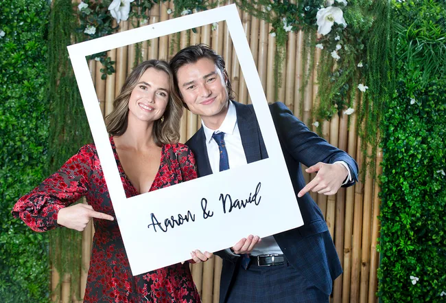 Two people smiling, holding a frame with "Aaron & David" written on it, standing in front of a foliage backdrop.