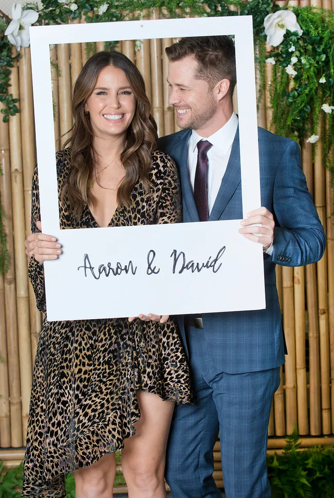 Couple smiling while holding a "Aaron & David" frame, standing in front of a bamboo and floral backdrop.