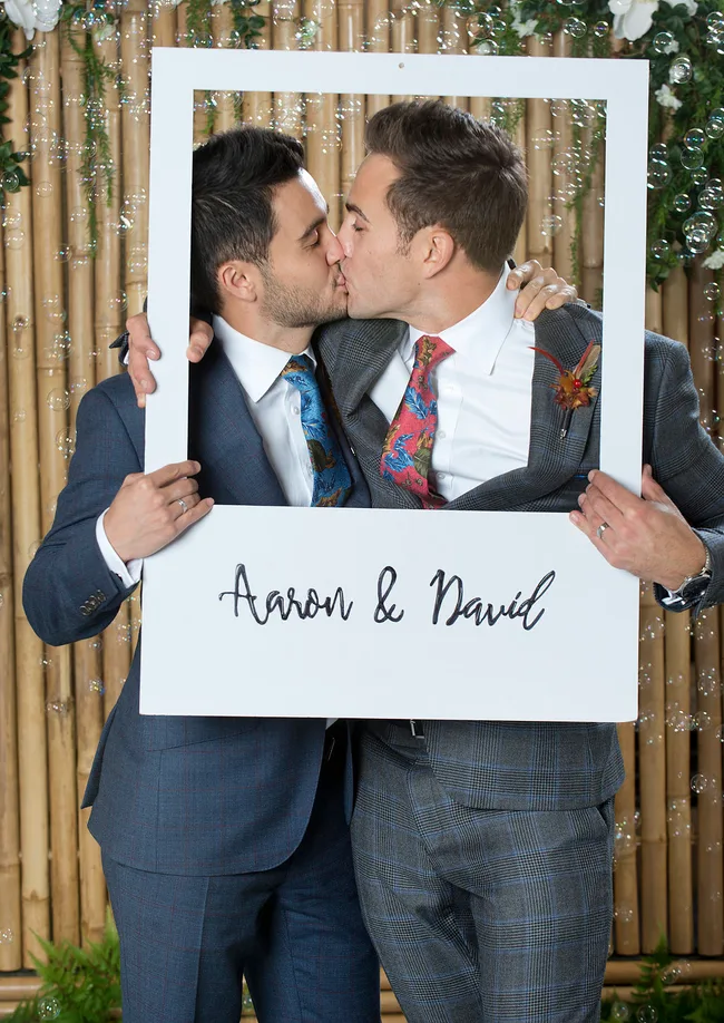 Two men in suits kissing, framed by a sign reading "Aaron & David" with a floral and bamboo background.
