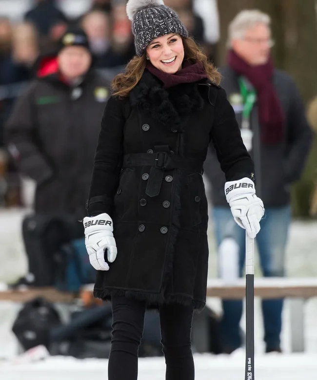 A person in winter attire smiles, wearing a knit hat and holding a hockey stick, with spectators in the background.