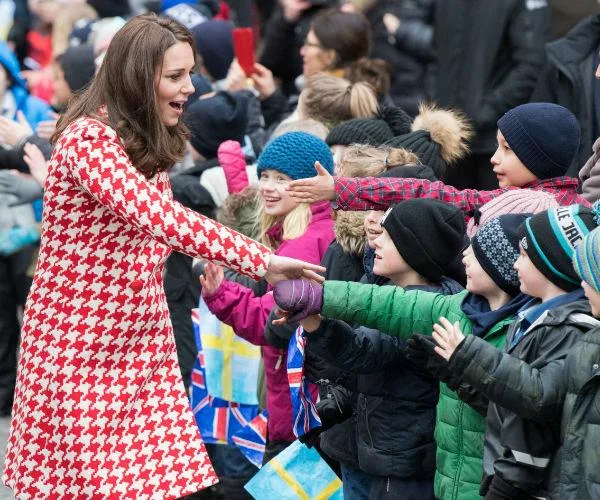 A woman in a red and white coat greets excited children holding flags in an outdoor crowd.