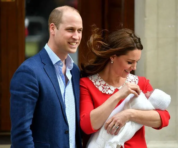 A couple smiling and looking at a newborn baby wrapped in a white blanket, standing outside a building.