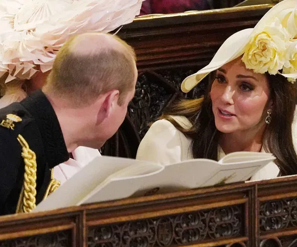 A man and woman in formal attire converse in a seated, ornate setting. Woman wears a large cream hat with yellow flowers.