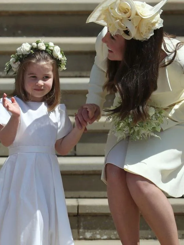 Woman in light outfit holding hands with a young girl in white dress and flower crown on steps, both smiling.