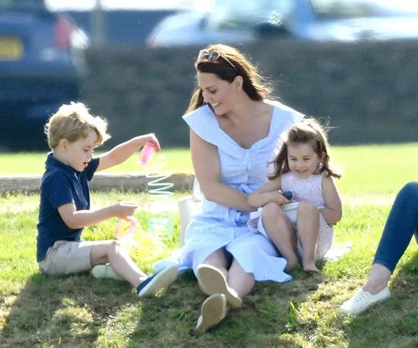 A mother and two children sit on grass; boy plays with a toy slinky, and the girl smiles, bright sunny day.
