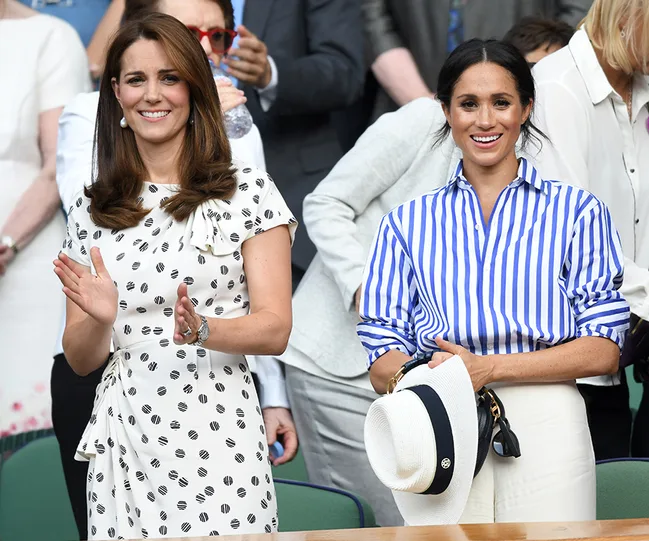 Two women standing and smiling, one clapping. The one on the left wears a polka dot dress, and the right one wears a striped shirt.