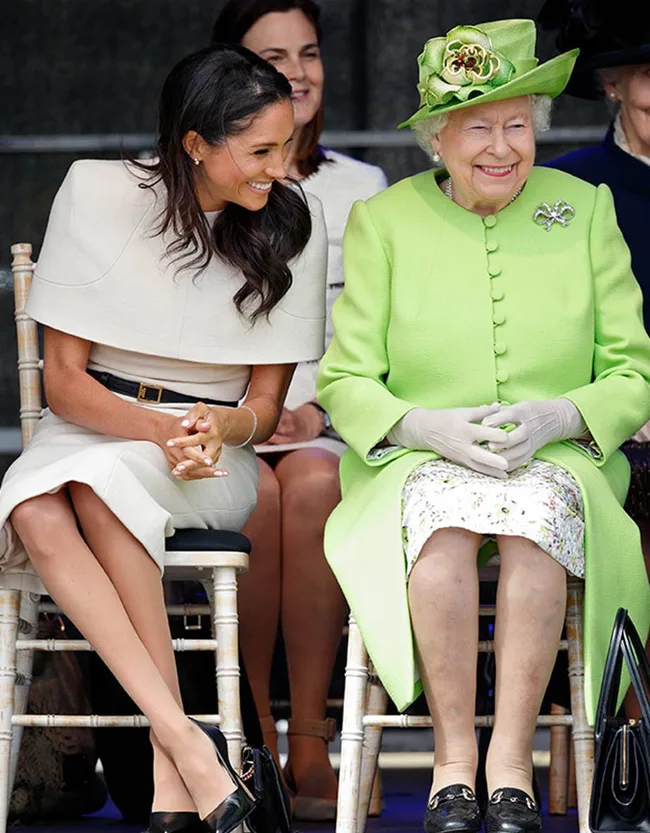 Two women seated, one in a light outfit and the other in a bright green suit and hat, smiling and conversing together.