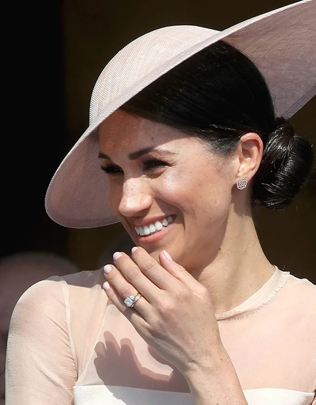 A person wearing a light pink hat and earrings, smiling with their hand near their face.