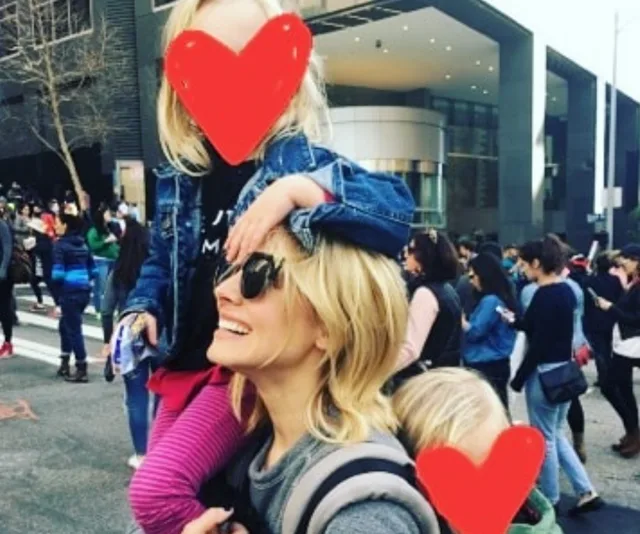 A smiling woman with sunglasses carries two children, faces covered with hearts, in a busy outdoor area.