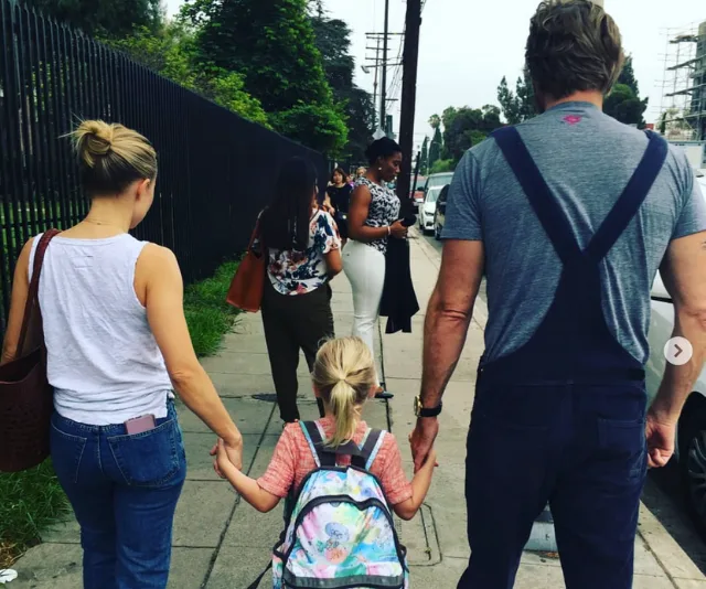 A man and woman holding a child's hands, walking on a sidewalk, child wearing a colorful backpack.
