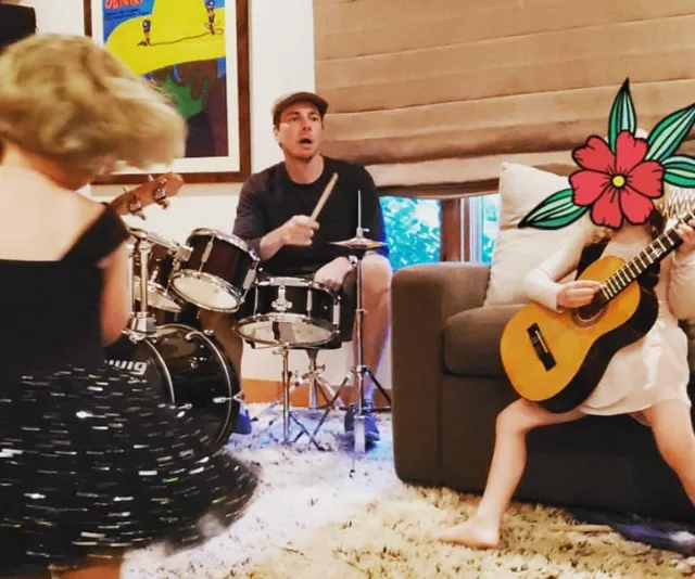 Man playing drums, child playing guitar, another child dancing in a room with a rug and artwork on the wall.