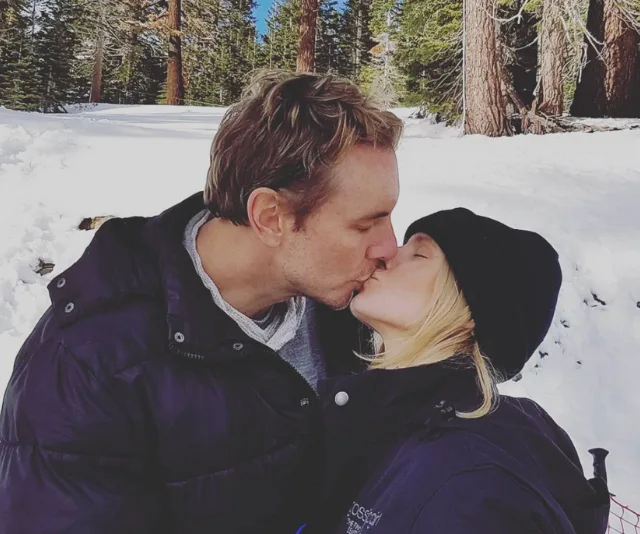 Couple kissing in a snowy forest, both wearing winter jackets and the woman wearing a black hat.