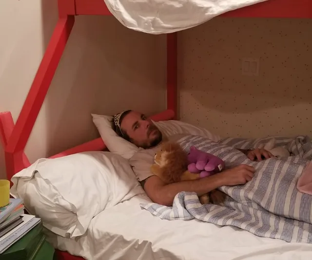 A man with a tiara lies in a bed under a striped blanket, holding plush toys.