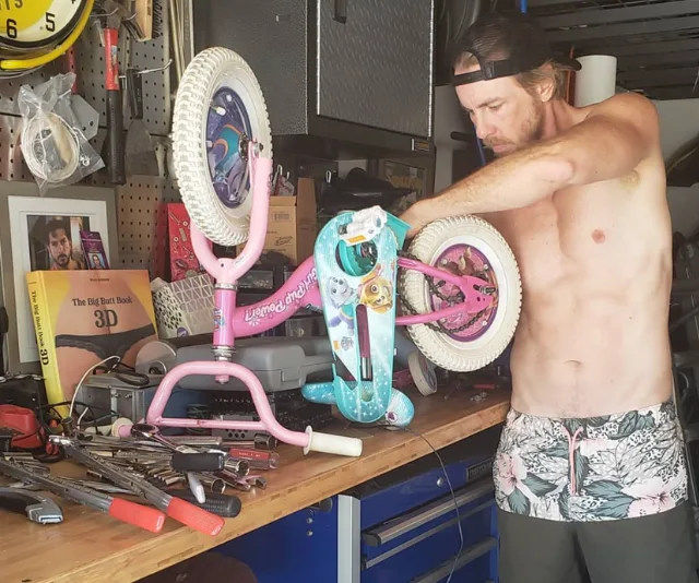 Man in a cap works shirtless on a child's bike in a garage, surrounded by tools and a book titled "The Big Butt Book."