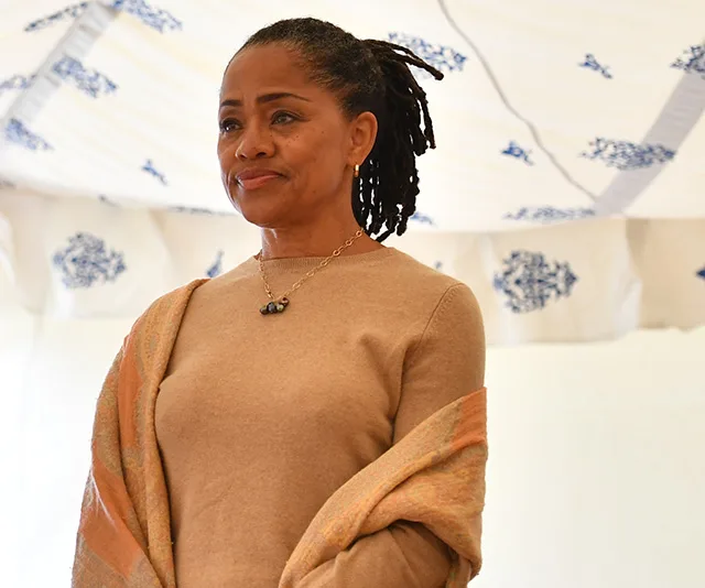 A woman with braided hair and a shawl stands under a patterned canopy, looking thoughtful.