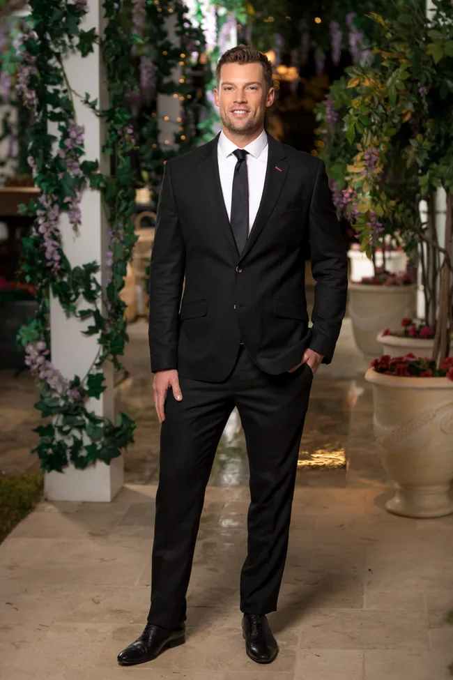 Man in a black suit and tie standing in a garden with greenery and flowers.