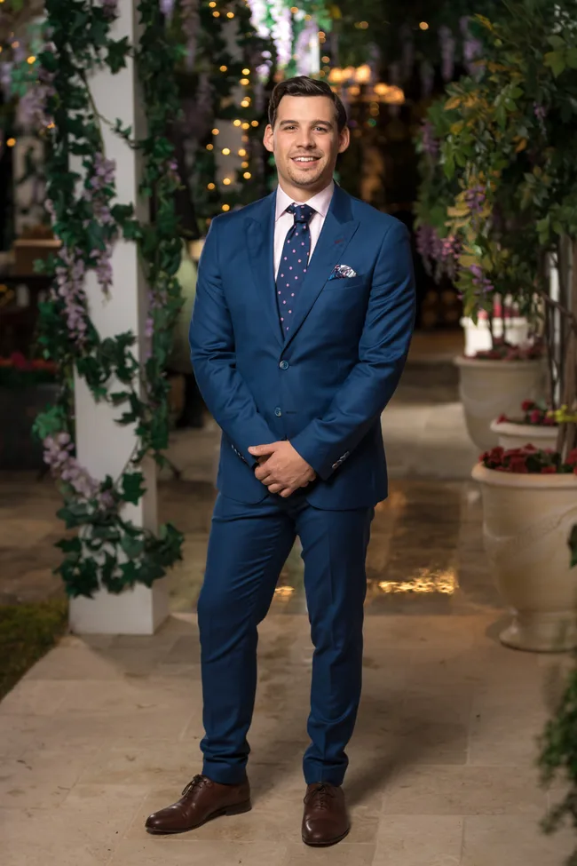 Man in a blue suit with a pink shirt and polka dot tie, standing in a garden setting with greenery and lights.