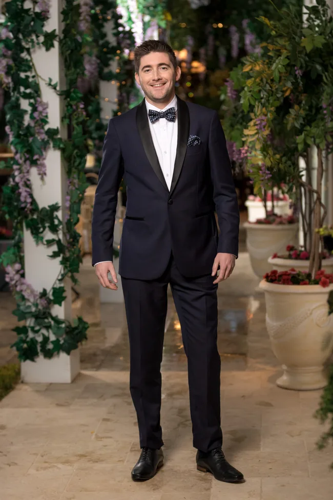 Man in a dark suit and bow tie standing in a garden setting with flowers and greenery around.