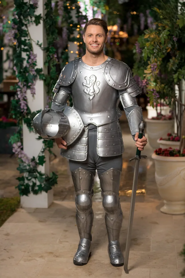 A man in shining knight armor holds a sword, standing on a patio decorated with greenery and flowers.