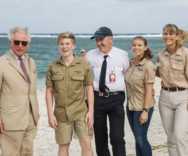 Group of five people, including a man in a tan suit and others in uniforms, smiling on a beach with ocean in the background.