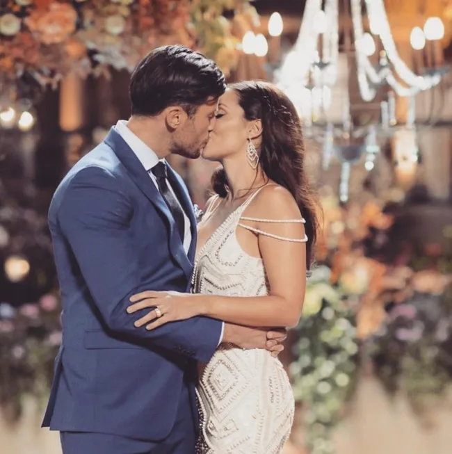 A couple kisses at their wedding, the groom in a blue suit and the bride in a white gown, under a chandelier.