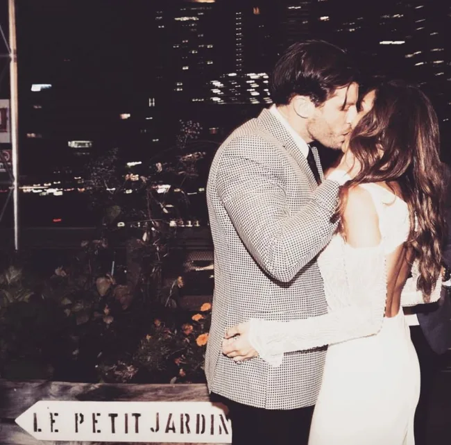 A couple kissing at night in formal attire with "Le Petit Jardin" sign and city lights in the background.