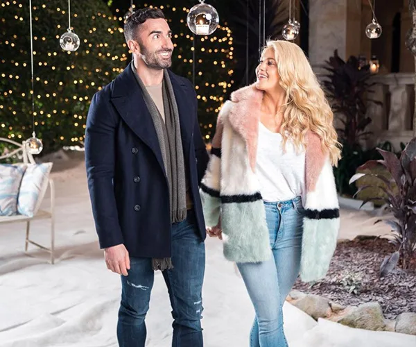 A couple walks hand in hand, smiling, outdoors with string lights in the background.