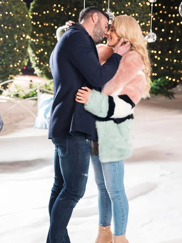 A couple embraces in a snowy winter setting with festive lights in the background.