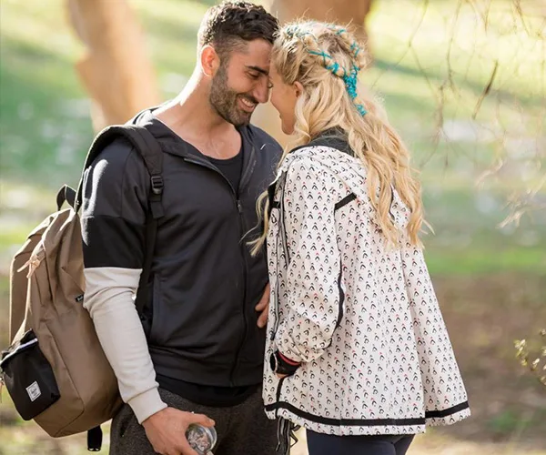 A man and woman stand close, smiling in a park. He carries a backpack; she has braided hair with blue flowers.