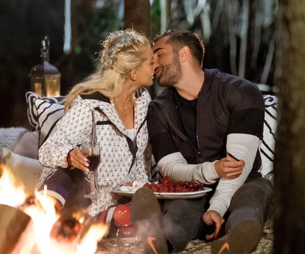 Couple sitting by a campfire, holding wine, leaning in for a kiss, with a plate of berries on their lap.