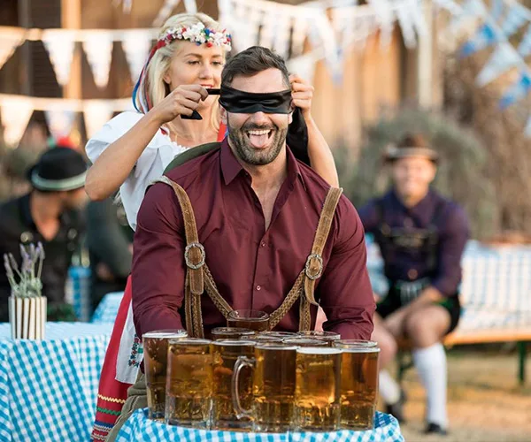 Man in lederhosen with blindfold, sitting in front of beer mugs at a festival; woman behind adjusts the blindfold.