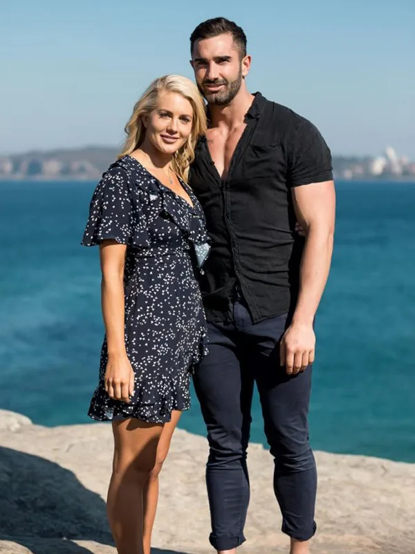 A couple stands together by the sea, smiling on a sunny day.
