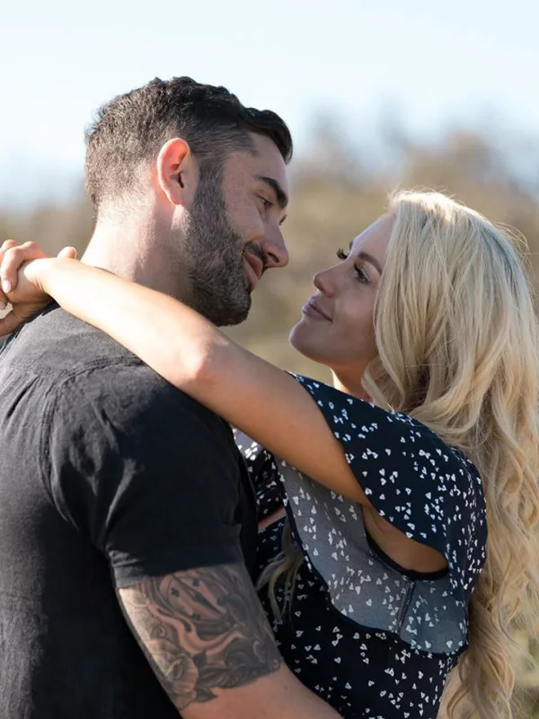 A couple embraces outdoors, smiling at each other with affection; the man has a tattoo on his arm.