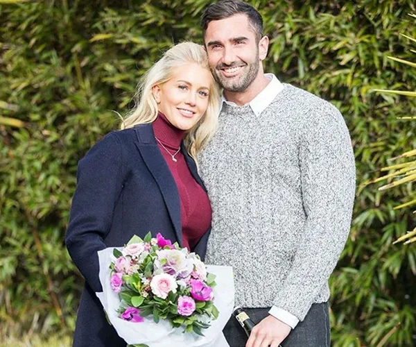 A smiling couple outdoors, woman holding a bouquet of flowers, standing close together in front of greenery.