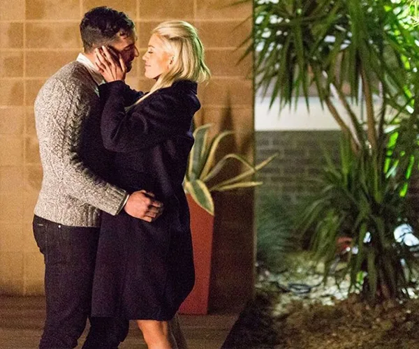 A couple embraces and kisses outdoors at night, surrounded by potted plants and a brick wall background.