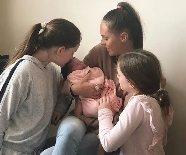 A woman holds a baby wrapped in a pink blanket, with two young girls looking on lovingly.