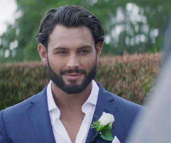 Man in a blue suit with a white shirt and boutonnière, smiling outdoors.
