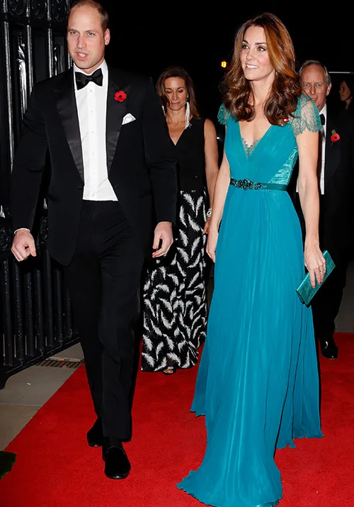 A man in a tuxedo and a woman in a teal gown walk on a red carpet, attended by others.