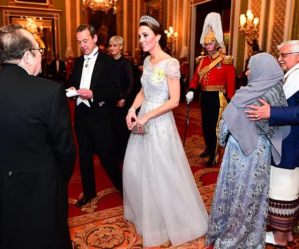 Gowned woman with tiara in formal gathering; ornate hall, guests in evening attire.