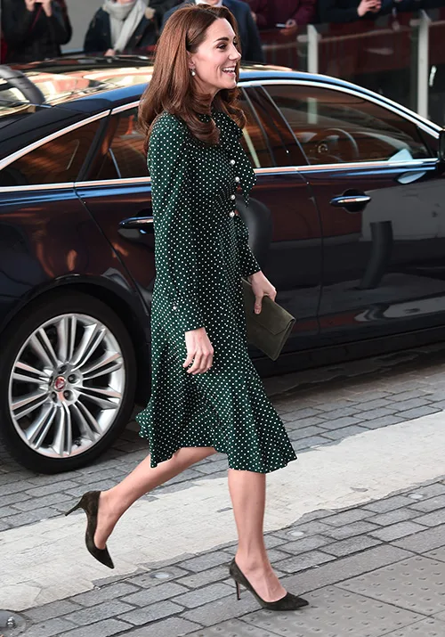 A woman in a green polka dot dress walks beside a black car on a cobblestone path, holding a clutch.
