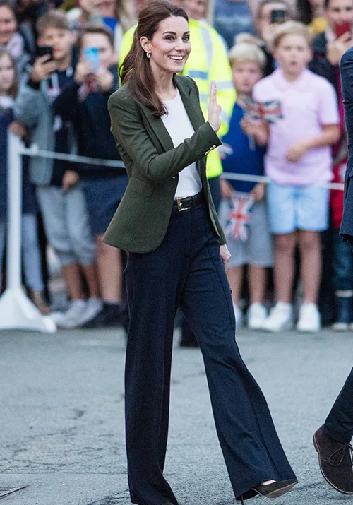 A woman in a green blazer waves to a crowd while walking, with people capturing the moment on their phones.