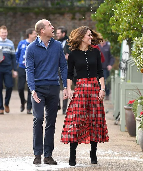 Couple walking outside, man in blue sweater and woman in black top with red plaid skirt, smiling at each other.