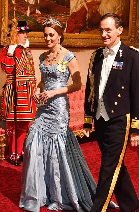 A woman in a light blue gown and tiara walks with a man in military attire; a uniformed guard stands behind them.