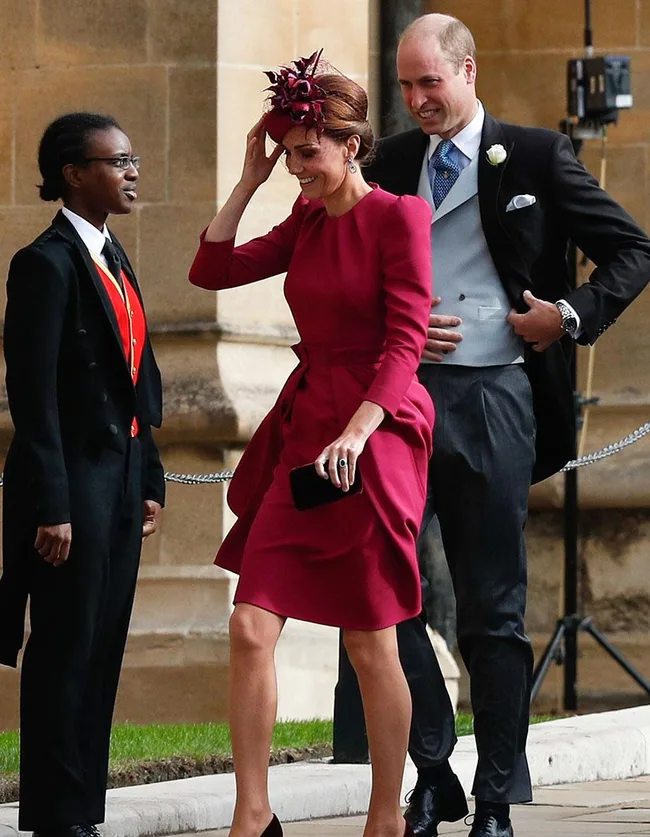 Two people dressed formally, with a woman in a red dress and hat, smiling outside a building.