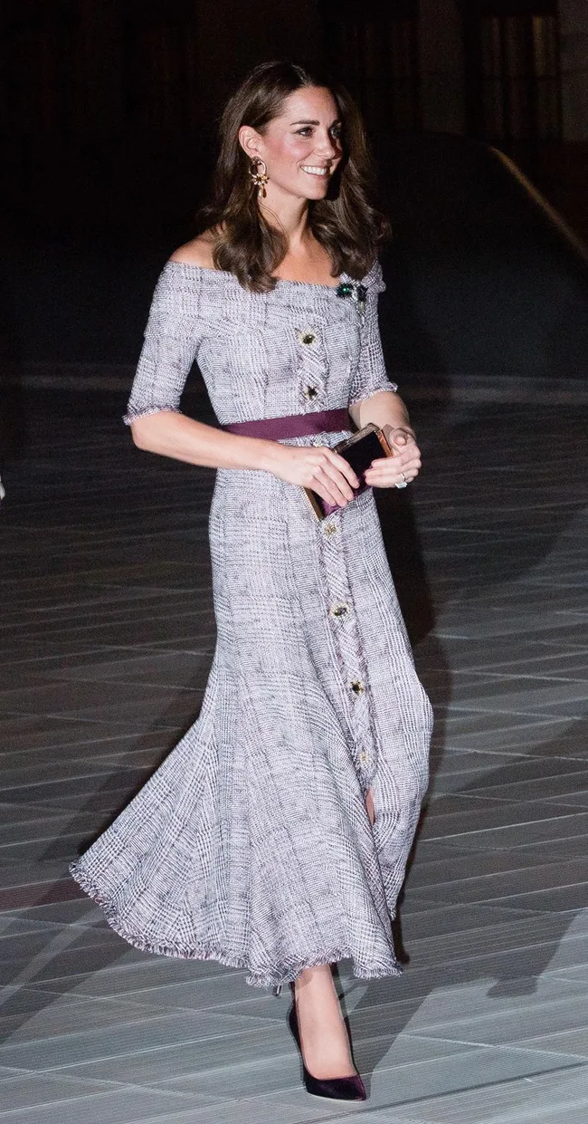 A woman in an off-shoulder grey checkered dress and heels, smiling while holding a clutch.