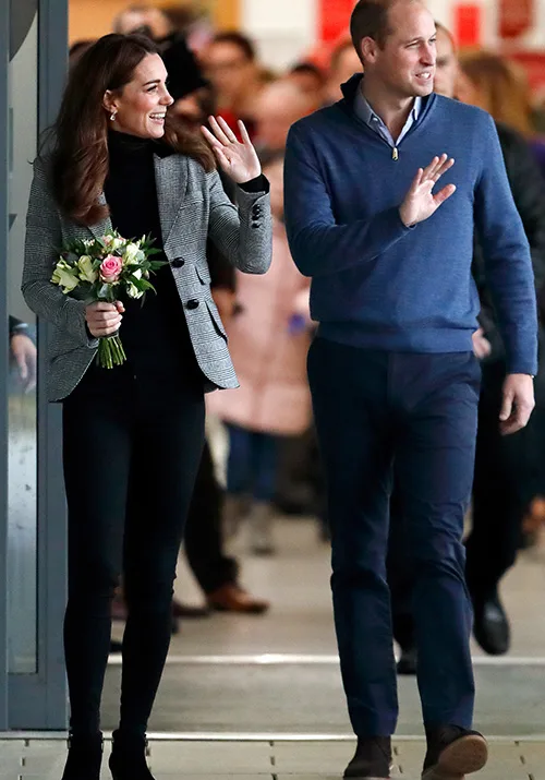 A woman and man wave while walking indoors; the woman holds a bouquet of flowers.
